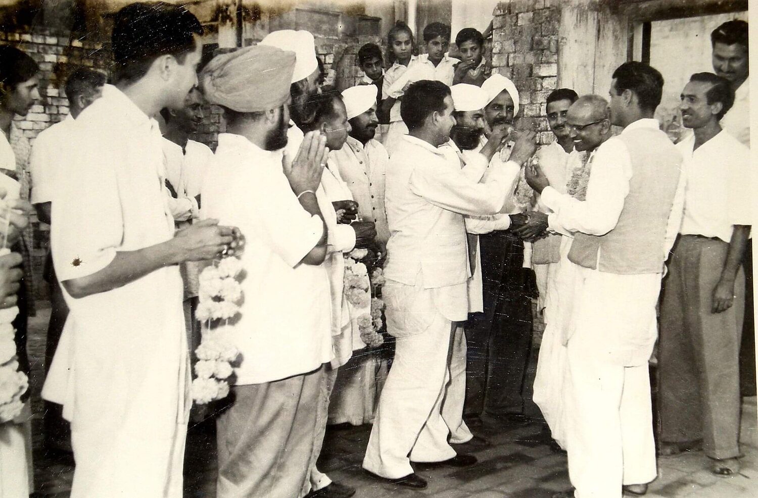 A crowd of people garlanding Batukeshwar Dutt at an outdoor gathering, with Dutt standing at the right in a white kurta.